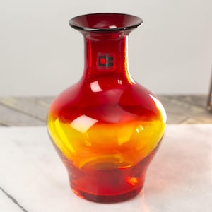 May include: A red and yellow glass vase with a rounded body and a narrow neck. The vase has a black label with a red circle and a black square.