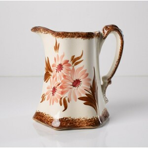 May include: A white ceramic pitcher with a brown handle and a floral design featuring pink and brown flowers. The pitcher has a brown rim and base.