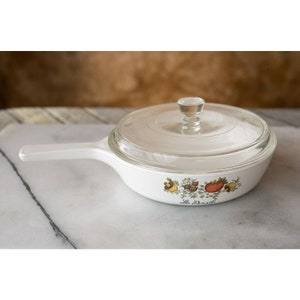 May include: White ceramic skillet with a glass lid and a white handle. The skillet has a floral design with a tomato, herbs, and spices. The text "Le  D'orsi" is printed on the bottom of the skillet.