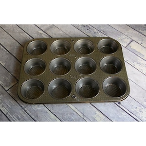 May include: A vintage metal muffin tin with 12 individual cups. The tin is dark gray and has a textured surface.