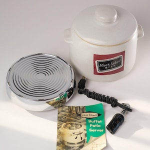 May include: A white ceramic crock pot with a lid and a black and red label that says "Black Label Beer". The crock pot is sitting on a silver electric warming base with a black cord. There is also a green and white instruction booklet for a "Buffet Patio Server" and a black electrical plug.