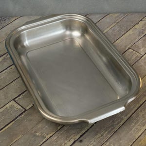 May include: A rectangular stainless steel baking pan with rounded corners and two side handles. The pan has a shiny, reflective surface and sits on a wooden surface.