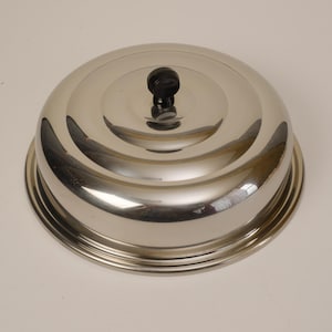 May include: A stainless steel food cover with a black knob handle. The cover is dome-shaped and sits on a matching circular base. The reflective surface of the cover creates a mirrored effect.
