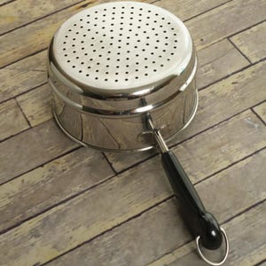 May include: A stainless steel colander with a cylindrical body and a flat, perforated top surface. The kitchen tool features a single, long, black handle attached to the side with a metal bracket and a small circular loop at the end for hanging.