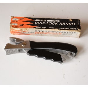 May include: A silver and black Anchor Hocking Grip-Lock Handle for cookware. The handle has a black plastic grip and a silver metal locking mechanism. The box reads "Anchor Hocking Grip-Lock Handle" and "clicks open...clicks shut...one-hand control".