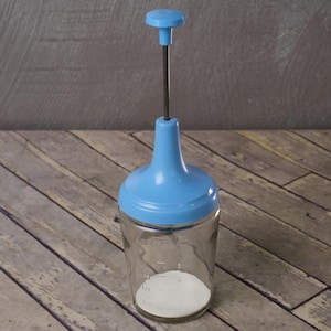 May include: A clear glass jar with a blue plastic top and plunger. The jar has measurement markings and contains a white substance. The plunger has a metal rod and a blue handle. The jar is on a wooden surface.