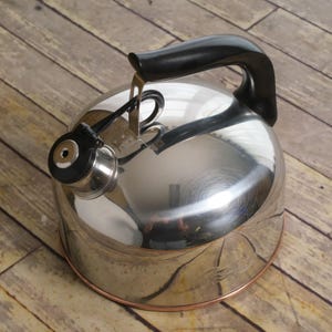May include: A polished stainless steel tea kettle with a rounded body, a black plastic handle, and a black whistle spout cover. The base of the kettle features a thin copper-colored trim, and the reflective surface shows a distorted environment.