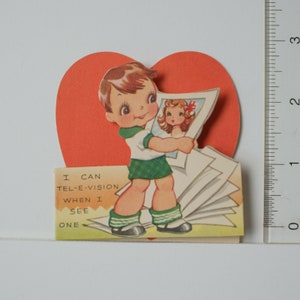 May include: A vintage Valentine's Day card featuring a cartoon boy holding a stack of photos, one of which shows a girl. The card has a red heart shape and the text "I can tel-e-vision when I see one".