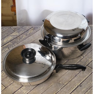 May include: Stainless steel cookware set with a frying pan, lid, and a multi-tiered pot with a lid. The frying pan has a black handle and a lid with a black knob. The pots are shiny and reflect the light.