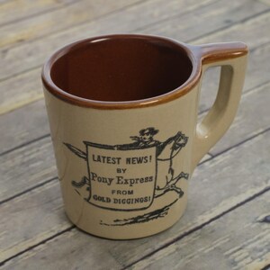 May include: A tan ceramic mug with a dark brown interior and rim. The exterior features a black graphic of a person on horseback carrying a sign that reads: LATEST NEWS! BY PONY EXPRESS FROM GOLD DIGGINGS! The mug has a thick, angular handle.