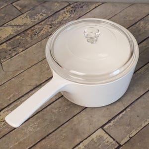 May include: White saucepan with a clear glass lid and a single handle. The cookware is made of a solid white material. The lid has a small, clear knob. The pan is sitting on a wooden surface.