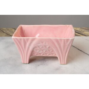 May include: A pink ceramic planter with a geometric design and four legs. The planter is made of glazed ceramic and has a smooth finish.