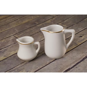 May include: Two white ceramic cream pitchers with gold trim. The pitchers are different sizes, with one being smaller than the other. Both have a handle and a spout for easy pouring. The pitchers are set against a wooden background.