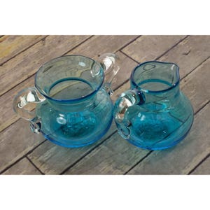May include: Two translucent turquoise glass pitchers with rounded bodies and clear glass handles. The larger pitcher features a wide mouth and a side handle, while the smaller pitcher has a distinct pouring spout and a matching clear glass handle.