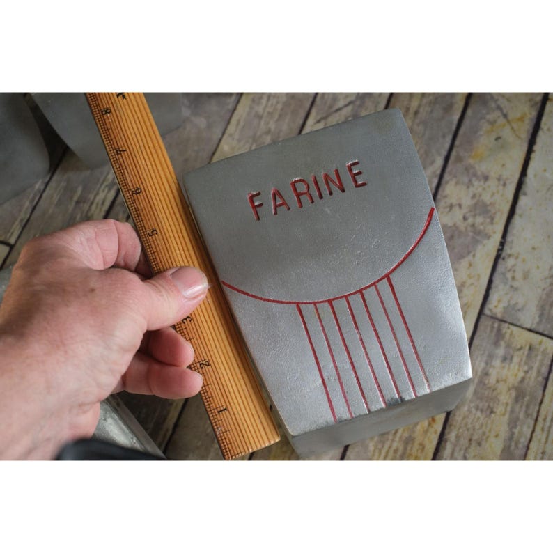 May include: A rectangular, silver-coloured container with the word "FARINE" in red lettering. The container has a curved red design and vertical red lines. A wooden ruler is next to the container.