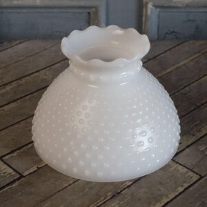 May include: A white hobnail glass lampshade with a ruffled top. The shade has a rounded, bell-like shape and is covered in small, raised dots. The lampshade is designed to diffuse light and add a vintage touch to any room.