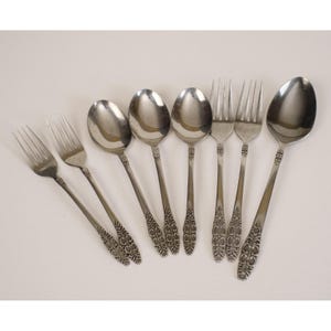 May include: A collection of stainless steel silverware, including spoons and forks, arranged on a white surface. The silverware features a decorative floral pattern on the handles.