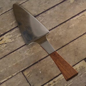 May include: A cake server with a triangular stainless steel blade and a straight wooden handle. The metal blade features a smooth, reflective surface and a serrated edge on one side, while the handle is made of brown wood with a visible grain.