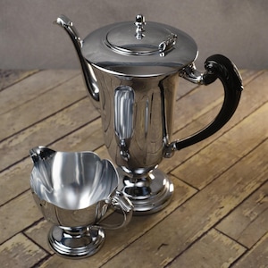 May include: A polished silver tea set, including a teapot with a black handle and a creamer. The teapot has a lid and sits on a pedestal base. The creamer has a scalloped rim and a small handle.