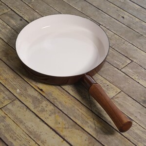 May include: A round frying pan with a glossy white interior and a dark brown exterior. The pan features a long, cylindrical wooden handle attached to the side, providing a contrast between the smooth enamel finish and the natural wood grain texture.