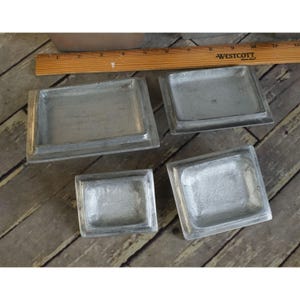 May include: Four small, rectangular, silver-coloured metal trays. The trays have a raised border and a recessed interior. A wooden ruler is visible in the background, with the word "WESTCOTT" printed on it.