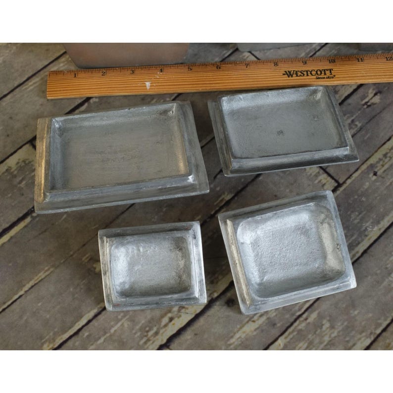 May include: Four small, rectangular, silver-coloured metal trays. The trays have a raised border and a recessed interior. A wooden ruler is visible in the background, with the word "WESTCOTT" printed on it.