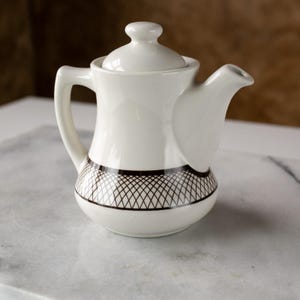 May include: A small, white ceramic teapot with a lid and handle. The base features a black and white crisscross pattern. The teapot is designed with a curved spout and a rounded body, suitable for serving tea.