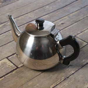 May include: A shiny, stainless steel teapot with a black handle and lid. The teapot has a curved spout and a hinged lid. The handle is black and curved. The teapot is sitting on a wooden surface.