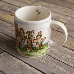 May include: White ceramic mug with a painted design of a wooden fence with vines and flowers. The flowers are pink, white, and orange.