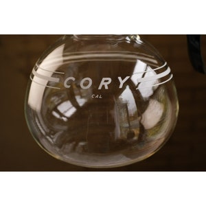 May include: A clear glass coffee pot with a white "CORY CAL" logo etched on the side. The pot has three white lines around the top.