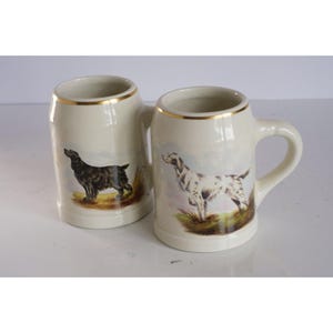 May include: Two ceramic beer steins with gold trim. One stein features a black and brown dog, the other a white and brown dog. Both dogs are depicted in a hunting scene.