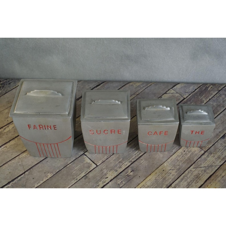 May include: A set of four vintage metal kitchen canisters. Each canister is square with a lid and a handle. The canisters are grey with red lettering that reads "FARINE", "SUCRE", "CAFE", and "THE".