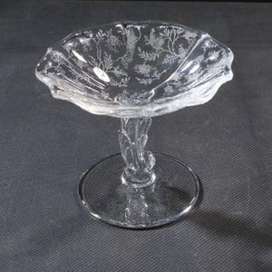 May include: A clear glass pedestal dish with a scalloped rim and etched floral design. The dish sits on a decorative stem and a round base. The glass is transparent, allowing light to pass through.