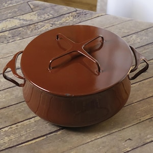 May include: A brown enameled cast iron Dutch oven with a matching lid. The lid features a cross-shaped handle. The pot has two side handles. The cookware is designed for stovetop or oven use.