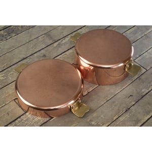 May include: Two copper pots with lids and brass handles. The pots are round and have a shiny finish. The handles are rectangular and attached to the sides of the pots. The pots are sitting on a wooden surface.