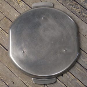 May include: A round, silver-colored metal baking pan with a smooth surface and three raised handles. The pan is designed for baking pies, cakes, or other desserts.