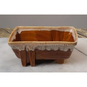 May include: A brown and white ceramic planter with a rectangular shape and four legs. The planter has a glazed finish with a white rim and a brown interior.