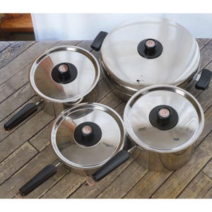 May include: A set of stainless steel cookware with lids. The set includes pots and pans of various sizes, each with a shiny, reflective surface. The lids have black handles and knobs. The cookware is arranged on a wooden surface.