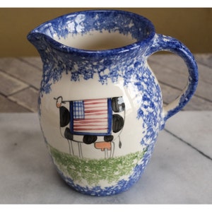 May include: A white ceramic pitcher with a blue and white speckled glaze. The pitcher features a hand-painted design of a black and white cow with an American flag on its back. The cow is standing on a green field.