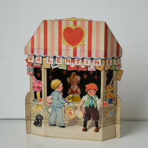 May include: A vintage Valentine's Day card featuring a paper pop-up stand with a red heart and the word "VALENTINES" on the front. The stand has a window with two children inside, one holding a hat and the other holding a book. There is a small dog in the front of the stand.