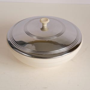 May include: A vintage, cream-colored serving bowl with a shiny, chrome-plated lid. The lid has a decorative, ridged design and a small, off-white knob. The bowl is round and appears to be made of ceramic or porcelain.