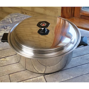 May include: A stainless steel cooking pot with a lid. The pot has a shiny, reflective surface and black handles. The lid features a black and red knob. The pot is sitting on a patterned surface.