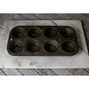 May include: A vintage, dark brown metal muffin tin with eight individual molds. The tin has a textured surface and is slightly worn.