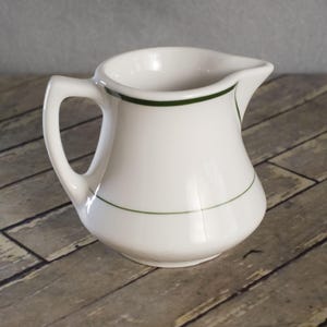 May include: A white ceramic pitcher with a curved body and a handle. The pitcher has a dark green trim around the top and a thin green line near the base. The pitcher is sitting on a wooden surface.