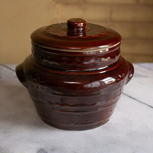 May include: A brown ceramic crock with a lid, featuring a decorative design of wavy lines and floral motifs. The crock has two handles on the sides.