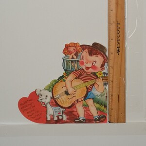 May include: Vintage Valentine's Day card featuring a cartoon boy playing a guitar, with a girl in the background. A white dog with a sign that says "To Be Mine" stands nearby. The card has a red heart with text.