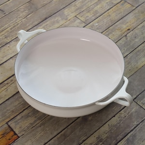 May include: A round, white enameled metal serving dish with a smooth interior and two curved handles on opposite sides. The rim of the dish features a thin, dark metallic edge, providing a clean contrast against the glossy white finish.