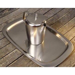 May include: A stainless steel teapot with a lid and a handle, sitting on a rectangular stainless steel tray. The teapot has a spout for pouring. The tray is slightly curved at the edges. The items are on a wooden surface.