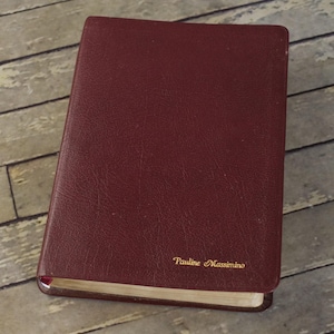 May include: A burgundy-colored book with a textured cover. The book has gold lettering that reads "Pauline Massimino" on the lower right corner. The pages are edged in gold. The book is closed and slightly angled.