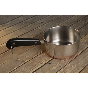 May include: A stainless steel saucepan with a copper-colored base and a black handle. The handle is attached with two rivets and has a loop for hanging. The pan is sitting on a wooden surface.
